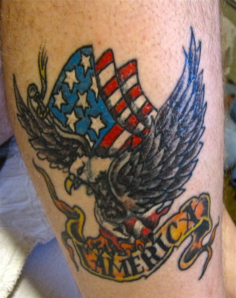 "A bold and colorful patriotic tattoo showcasing an American bald eagle soaring with the U.S. flag draped behind it. The tattoo highlights national pride and features a ribbon with the word 'America,' symbolizing strength and freedom.