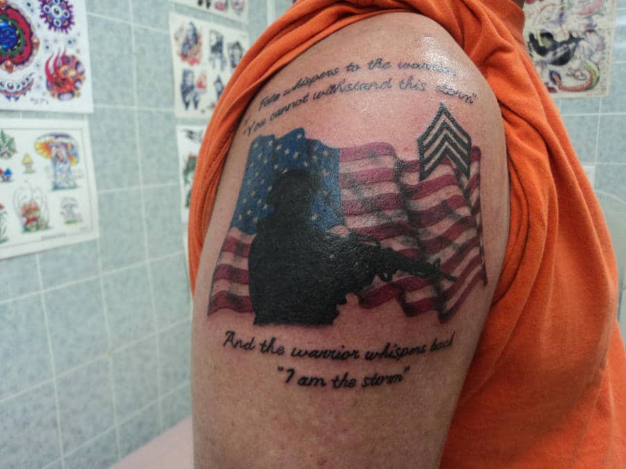 An army-themed tattoo for guys featuring a silhouette of a soldier holding a rifle, set against a waving American flag. Above the image, a quote reads, 'Fate whispers to the warrior, you cannot withstand this storm,' followed by 'And the warrior whispers back, I am the storm' below the image. The design is completed with military chevrons, emphasizing strength, courage, and patriotism.