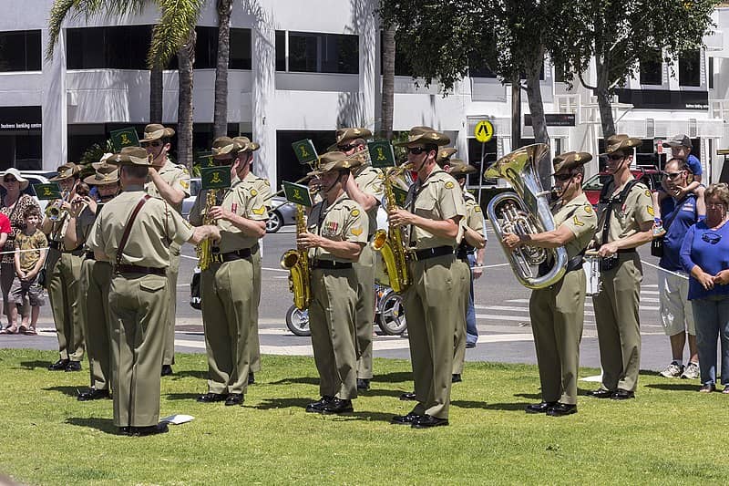 The Band of the Royal Australian Navy marching in a grand parade, performing with brass and percussion instruments as large crowds line the streets to watch. The band, dressed in formal naval uniforms, leads a procession that exemplifies the pride and tradition of the Royal Australian Navy.