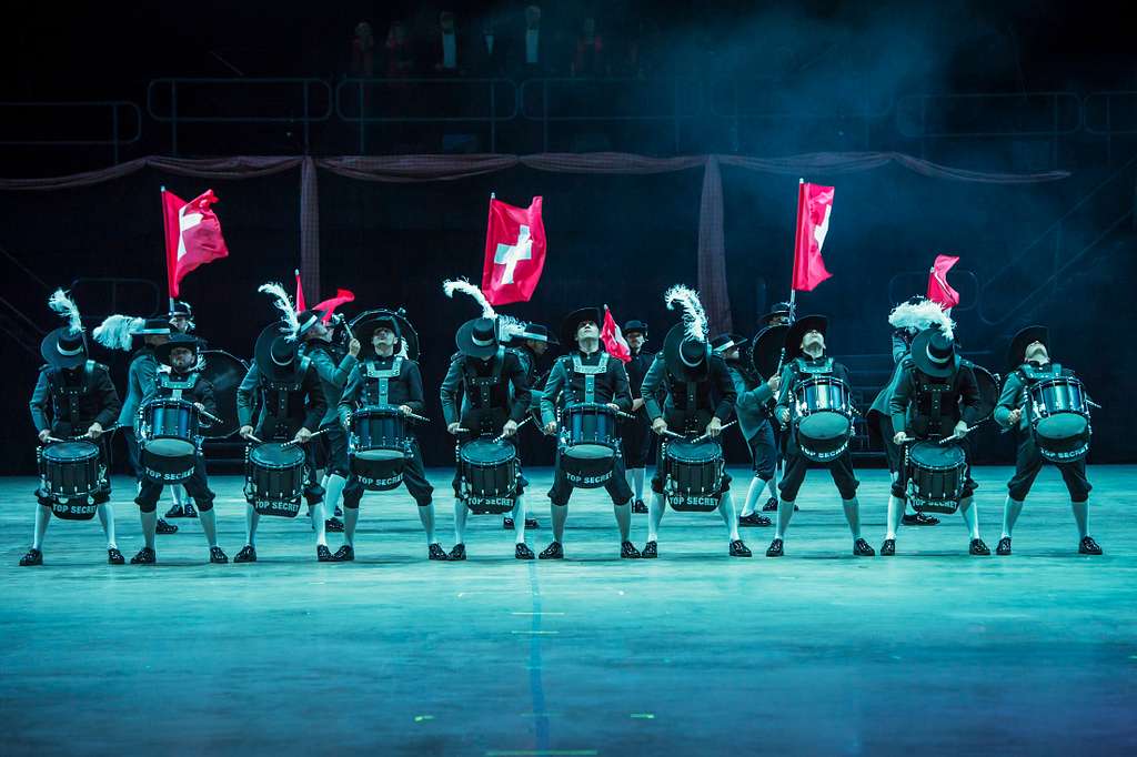 A dramatic performance at the Basel Tattoo featuring a drumline dressed in traditional attire, with performers waving Swiss flags under moody lighting. This scene captures the energy and precision often highlighted in a Basel Tattoo guide, offering insight into the event’s unique and dynamic performances.