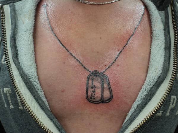 A realistic dog tag tattoo inked on the chest, designed to resemble military identification tags hanging from a chain around the neck. The tattoo is intricately detailed, giving the illusion of the dog tags resting naturally on the skin, symbolizing the wearer's connection to the military and honoring service or fallen comrades.