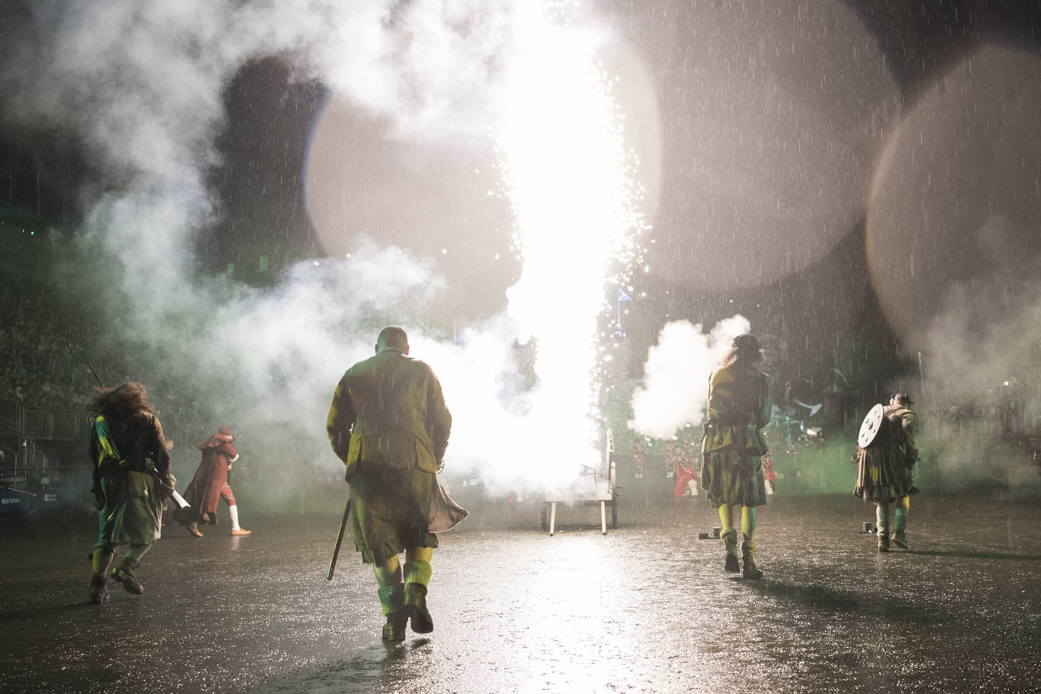 Performers dressed in traditional Scottish attire, including kilts, walking away from a dramatic explosion of light and smoke during a military tattoo. The scene captures the intensity and spectacle of the event, emphasizing the importance of timing your visit to experience the full duration of the show. The atmosphere is heightened by the rain, adding a dynamic element to the performance.