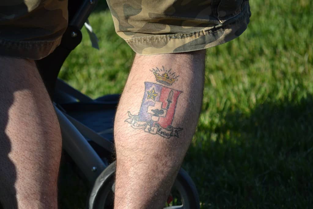 A military insignia tattoo on the calf, featuring a colorful emblem with a shield, a crown, and a scroll with text. The design is detailed and symbolizes the wearer's affiliation with a specific military unit or family heritage. The tattoo is set against a backdrop of camouflage shorts, emphasizing the military theme.