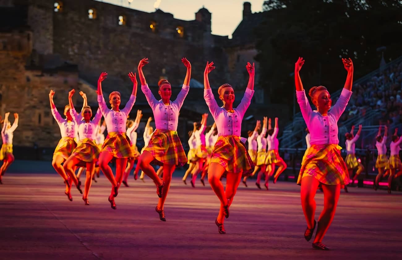 A vibrant group of dancers performing in colorful tartan skirts during a Military Tattoo event, set against the backdrop of a historic castle. This scene embodies the energy and cultural celebration expected at Military Tattoo Events in 2025.