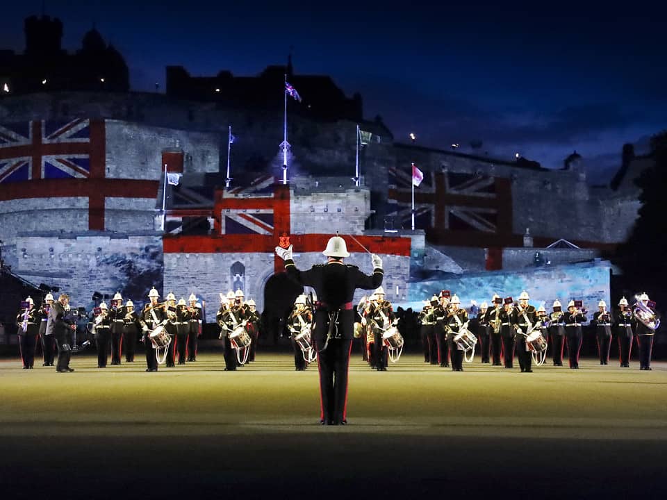 A military tattoo performance featuring a band of uniformed musicians playing at Edinburgh Castle, with the Union Jack projected onto the castle walls, creating a striking backdrop.