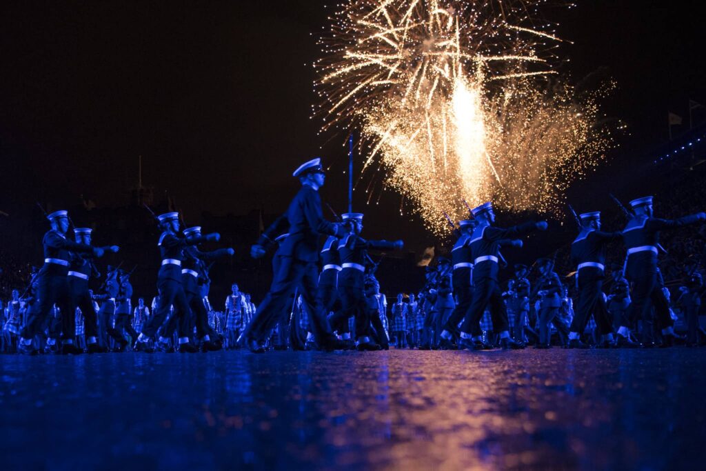 A military performance at a tattoo show under a nighttime sky with fireworks illuminating the background. The soldiers in uniform march in unison, bathed in blue light, while spectators watch from the stands, creating a dramatic and festive atmosphere.