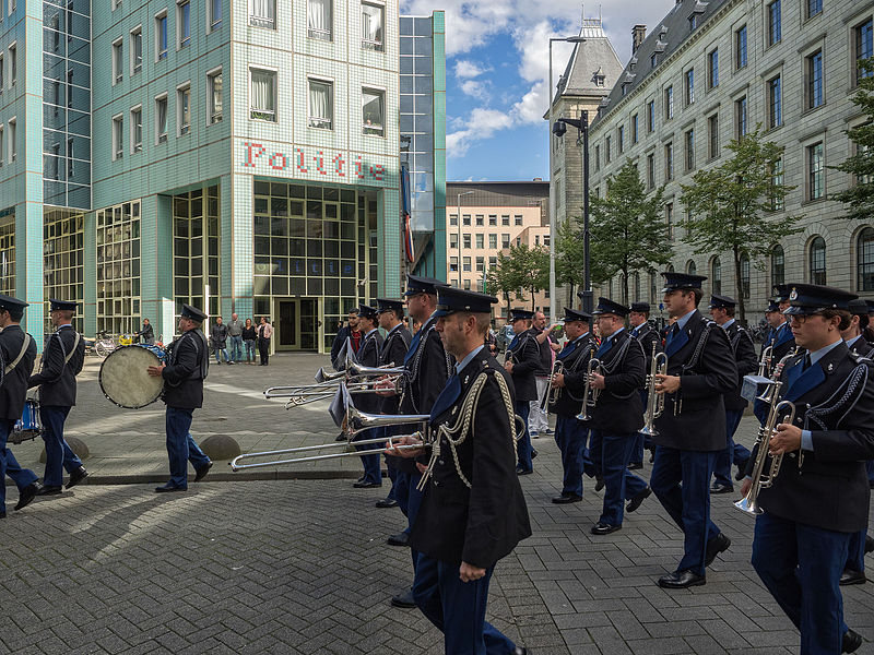 A military band dressed in formal uniforms marching through the streets of Rotterdam during the Rotterdam Military Tattoo, with musicians playing brass and percussion instruments in front of modern and historic buildings, showcasing the city's blend of tradition and modernity.