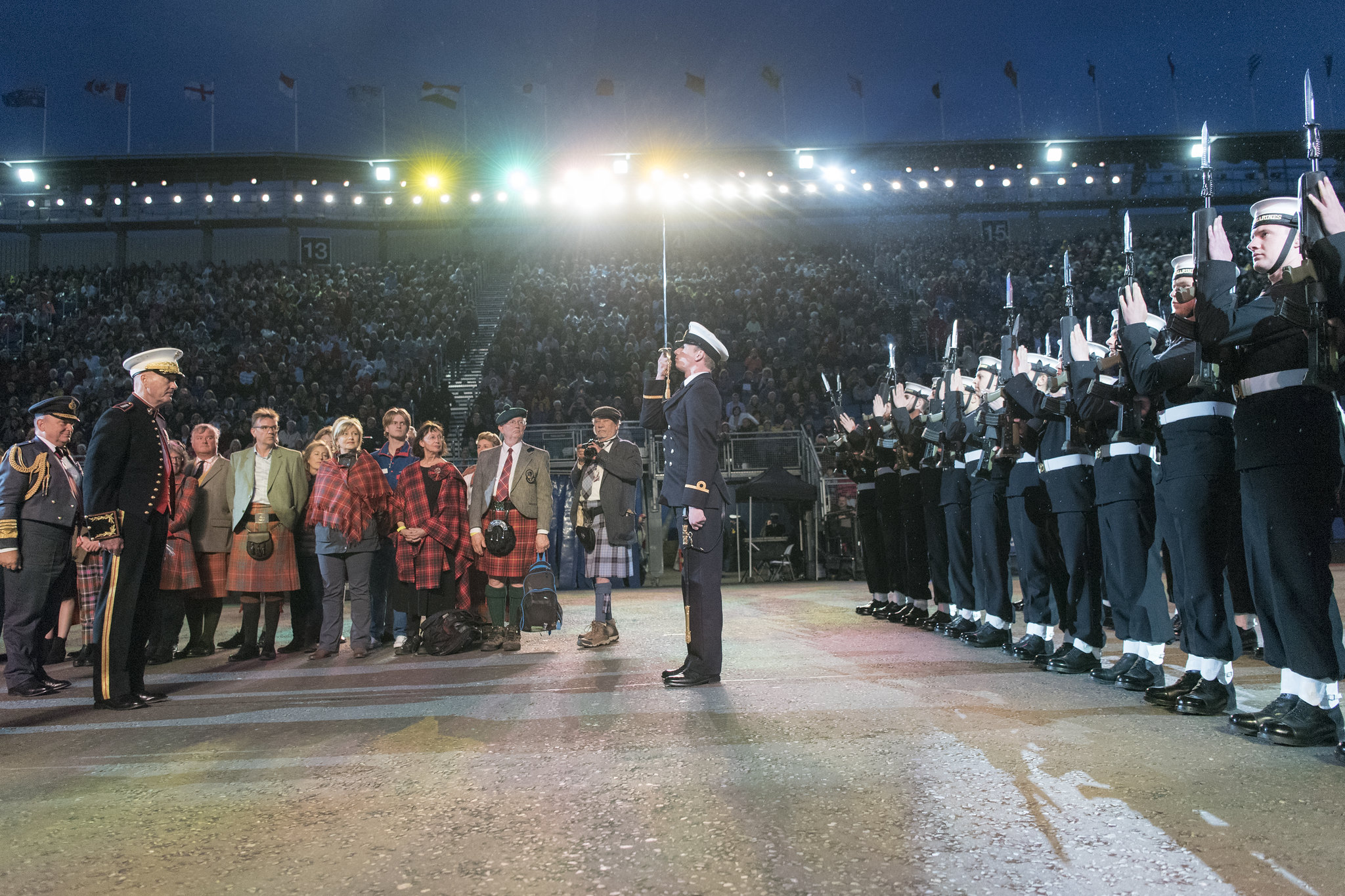 A historic scene at the Royal Edinburgh Military Tattoo, featuring military personnel in formal uniforms and a group of people in traditional Scottish attire. The event, held at Edinburgh Castle, is a significant part of the Royal Edinburgh Military Tattoo history. The grandstand is filled with spectators under bright lights, reflecting the ceremony's importance and tradition