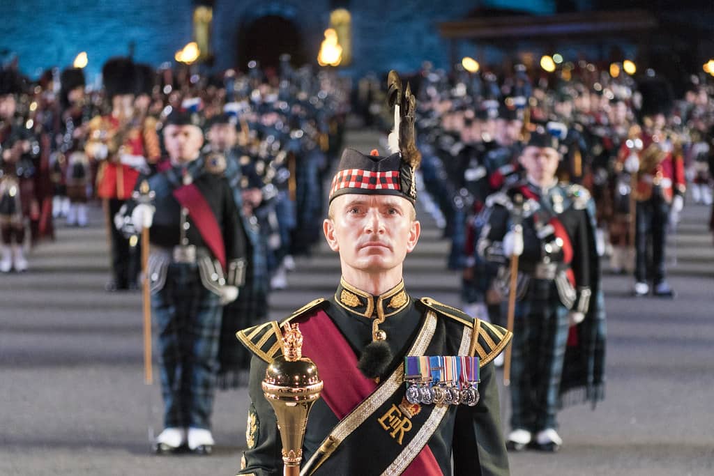 A ceremonial leader in full traditional military attire stands at the forefront of a formation during the Edinburgh Military Tattoo, with other uniformed participants in the background. The image captures the precision and grandeur of the event, offering a glimpse of what attendees can expect when learning how to get tickets for the Edinburgh Military Tattoo.