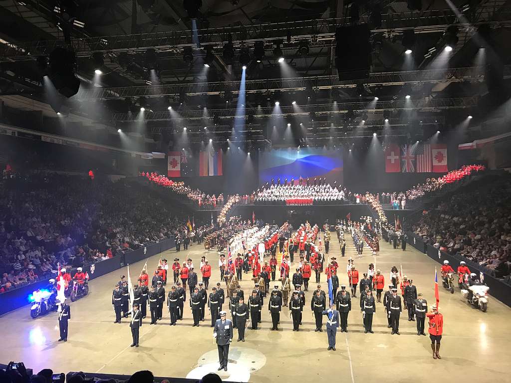 A grand procession of military bands and performers at the Royal Nova Scotia International Tattoo, with participants in various uniforms marching in formation under bright stage lights in an indoor arena, surrounded by an audience. The scene captures the international and ceremonial spirit of this prestigious event.