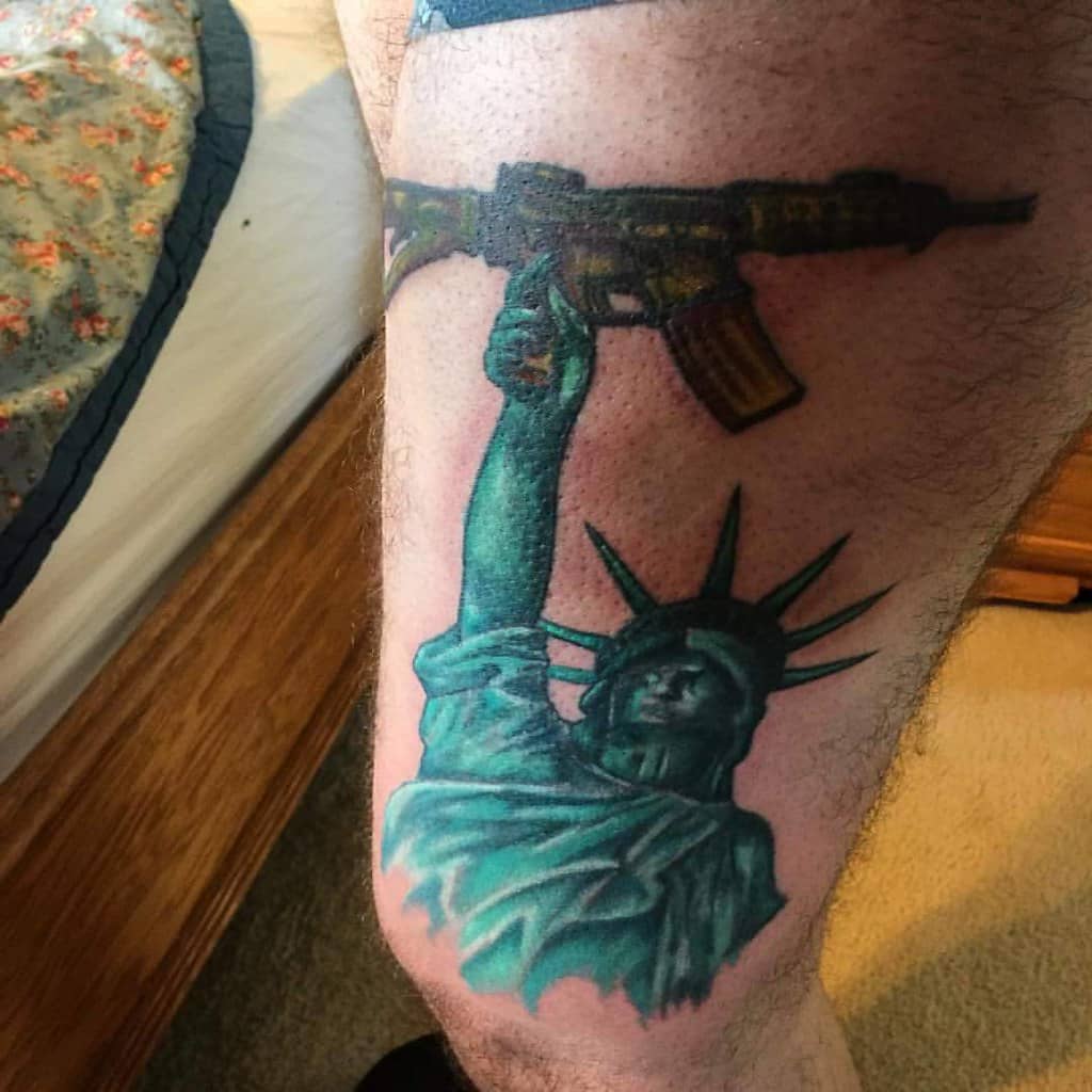 A bold tattoo featuring the Statue of Liberty holding up an assault rifle instead of the traditional torch. The design blends iconic American symbolism with a military theme, representing a fusion of liberty, strength, and defense. The detailed shading and vibrant colors make the tattoo stand out on the leg, conveying a powerful statement.