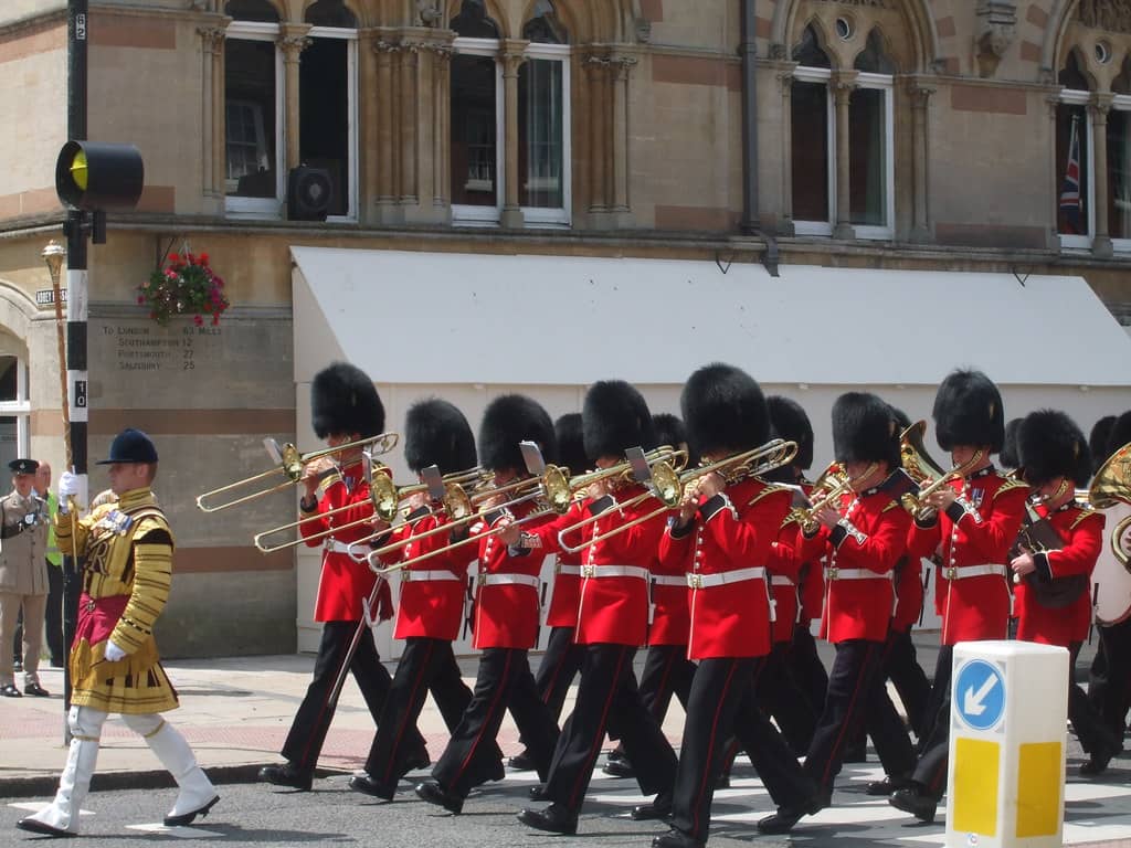 The Band of the Grenadier Guards, a famous military band, marching in formation through the streets in their iconic red uniforms and bearskin hats, performing with brass instruments under the direction of a band leader in ceremonial attire.