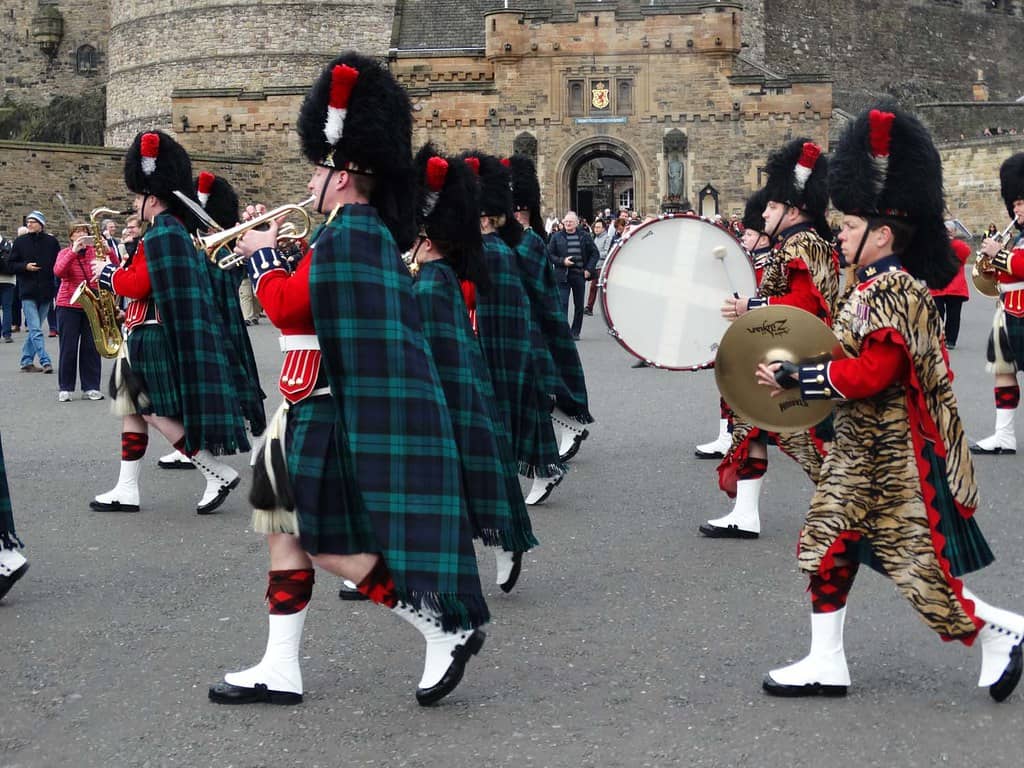 The Band of the Royal Regiment of Scotland, a famous military band, marching in full Highland dress with kilts, sporrans, and bearskin hats, playing brass and percussion instruments in front of a historic castle, with spectators watching the performance.