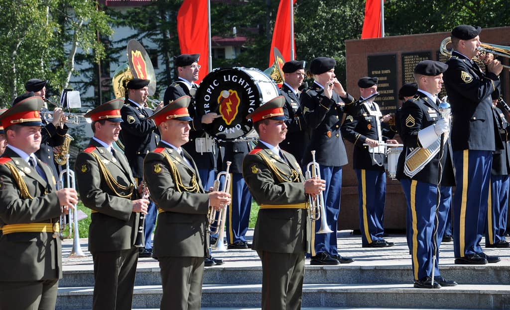 The Central Military Band of the Ministry of Defense of Russia, a famous military band, performing at a ceremonial event with musicians in green military uniforms and red caps standing in formation, alongside members of an allied military band in black and blue uniforms, playing brass and percussion instruments.