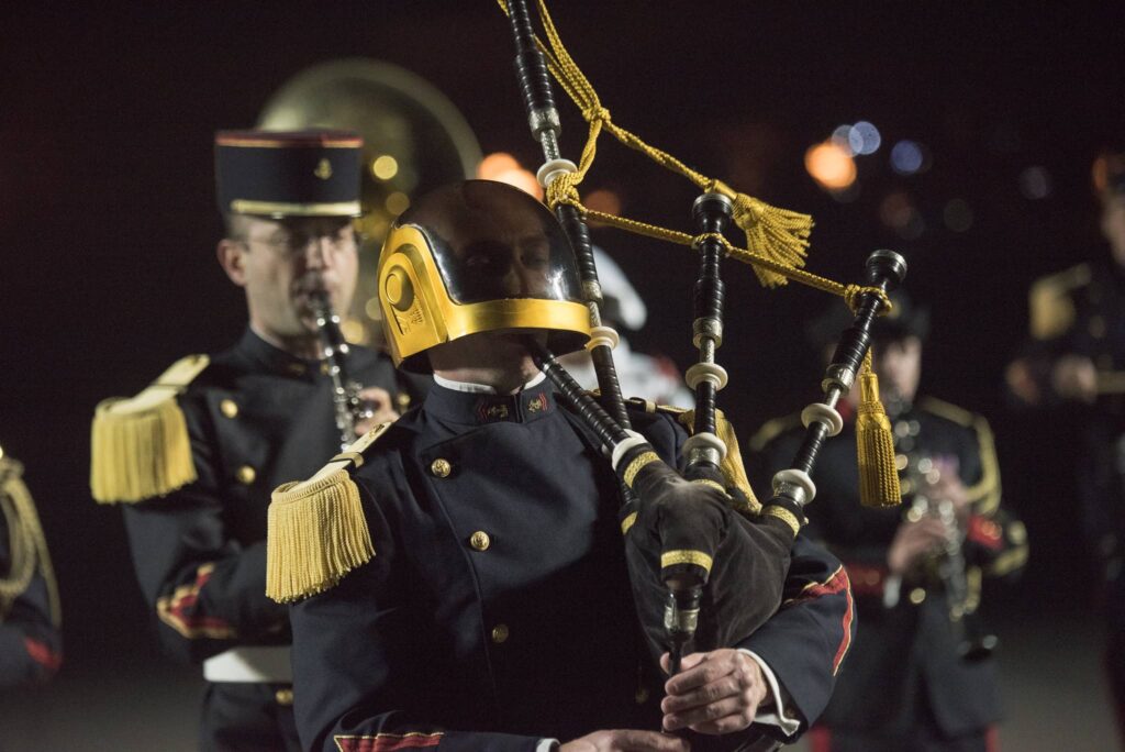 A close-up of a musician playing the bagpipes during a military tattoo performance, wearing a distinctive golden helmet and formal military uniform, with other band members in the background.