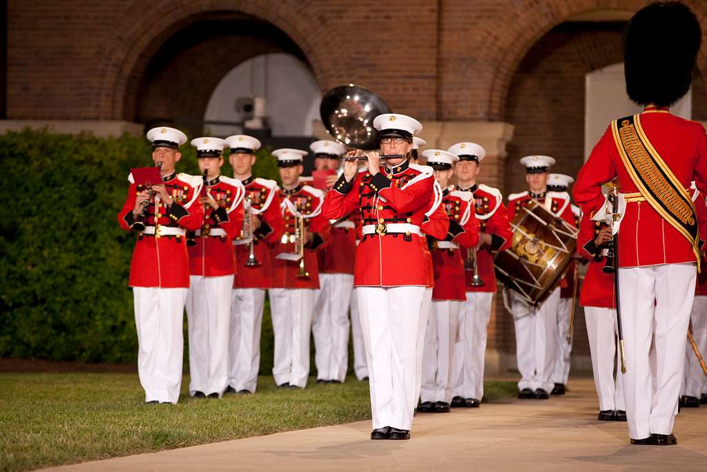 The United States Marine Band, a famous military band, performing in their iconic red and white uniforms with brass and percussion instruments during a ceremonial event in front of a historic building.