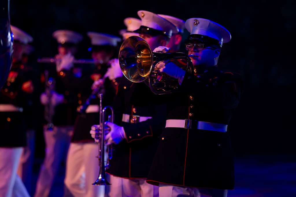 A musician in a United States Marine Corps uniform performing on a brass instrument during the Virginia International Tattoo, with fellow band members in the background under dramatic stage lighting. This image captures the precision and honor associated with this renowned military event.