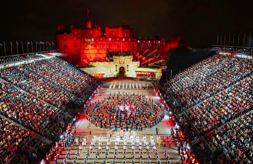 A breathtaking view of the Royal Edinburgh Military Tattoo, with massed military bands performing in front of a packed audience at Edinburgh Castle, illuminated in vibrant red lighting. The event showcases the precision and pageantry of international military bands, exemplifying what the Royal Edinburgh Military Tattoo is known for.