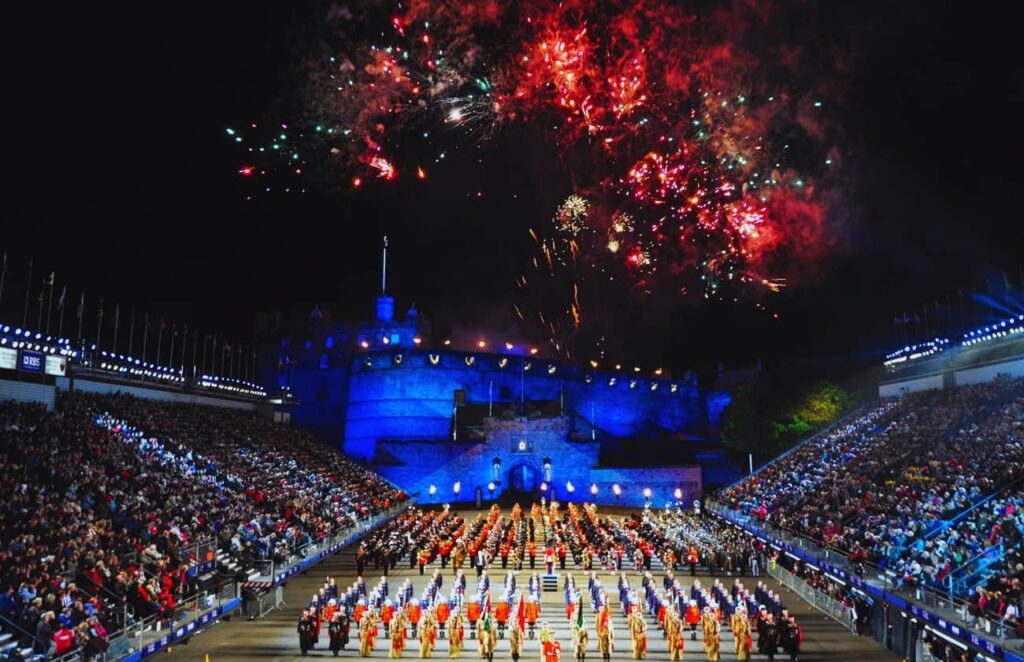 A stunning display at the Royal Edinburgh Military Tattoo in Scotland, with massed military bands in colorful uniforms performing in front of Edinburgh Castle, illuminated in blue, under a sky filled with fireworks. This image captures the grandeur and significance of what a military tattoo in Scotland entails.