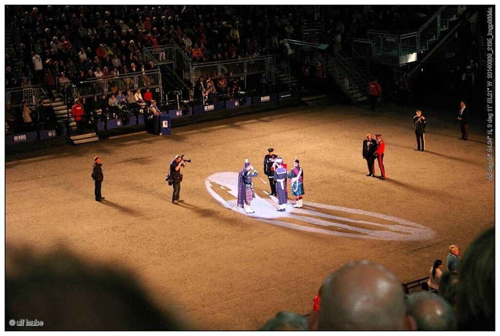 An evening performance at the Edinburgh Military Tattoo held at Edinburgh Castle, where the Edinburgh Military Tattoo is held. The image shows performers in traditional Scottish attire illuminated by a spotlight on the parade ground, with a large audience watching from the stands in the background.