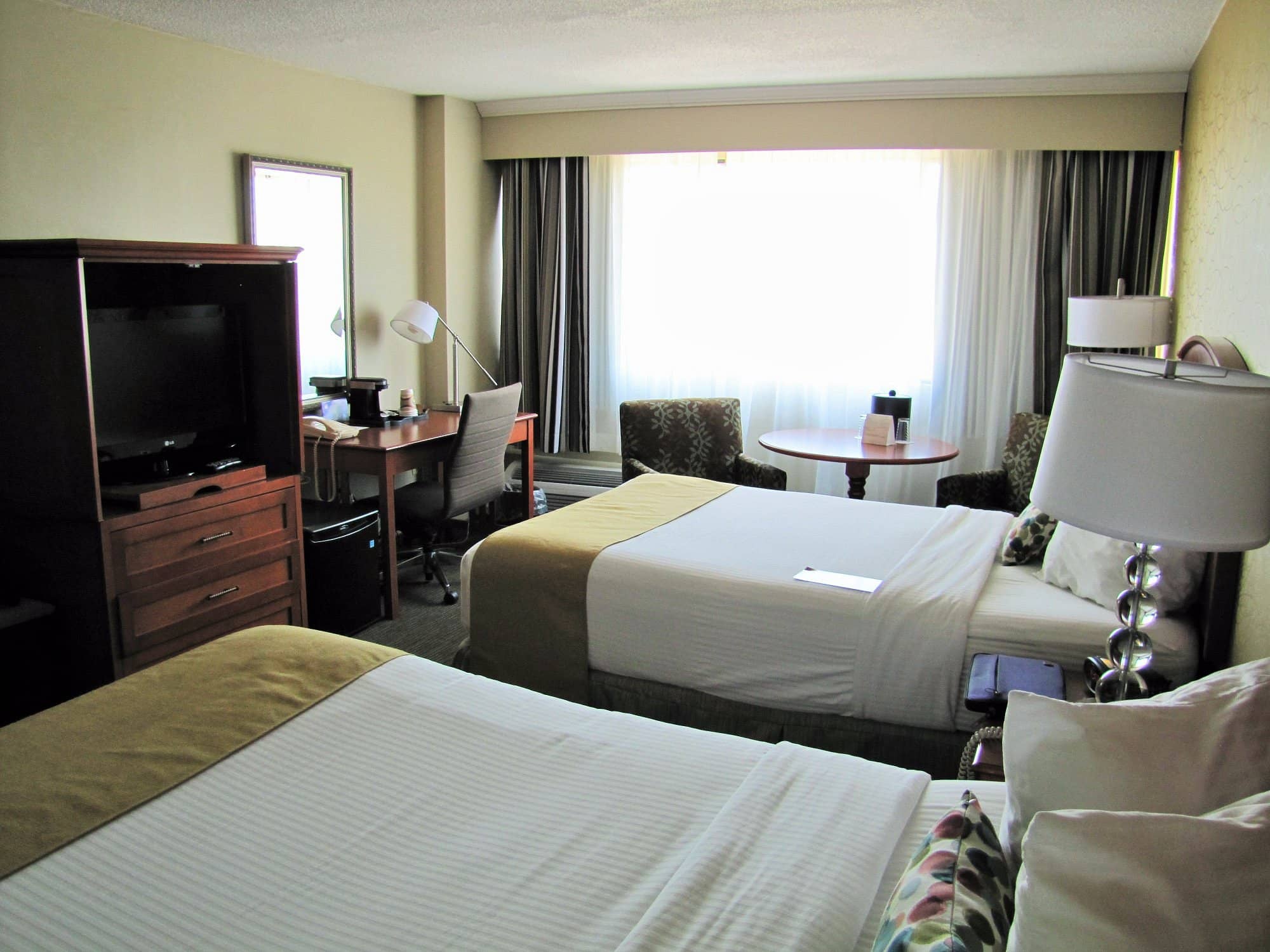 A bright and inviting room at Atlantica Hotel Halifax featuring two double beds with white linens a work desk a TV in a wooden cabinet and a small table with two chairs by the window