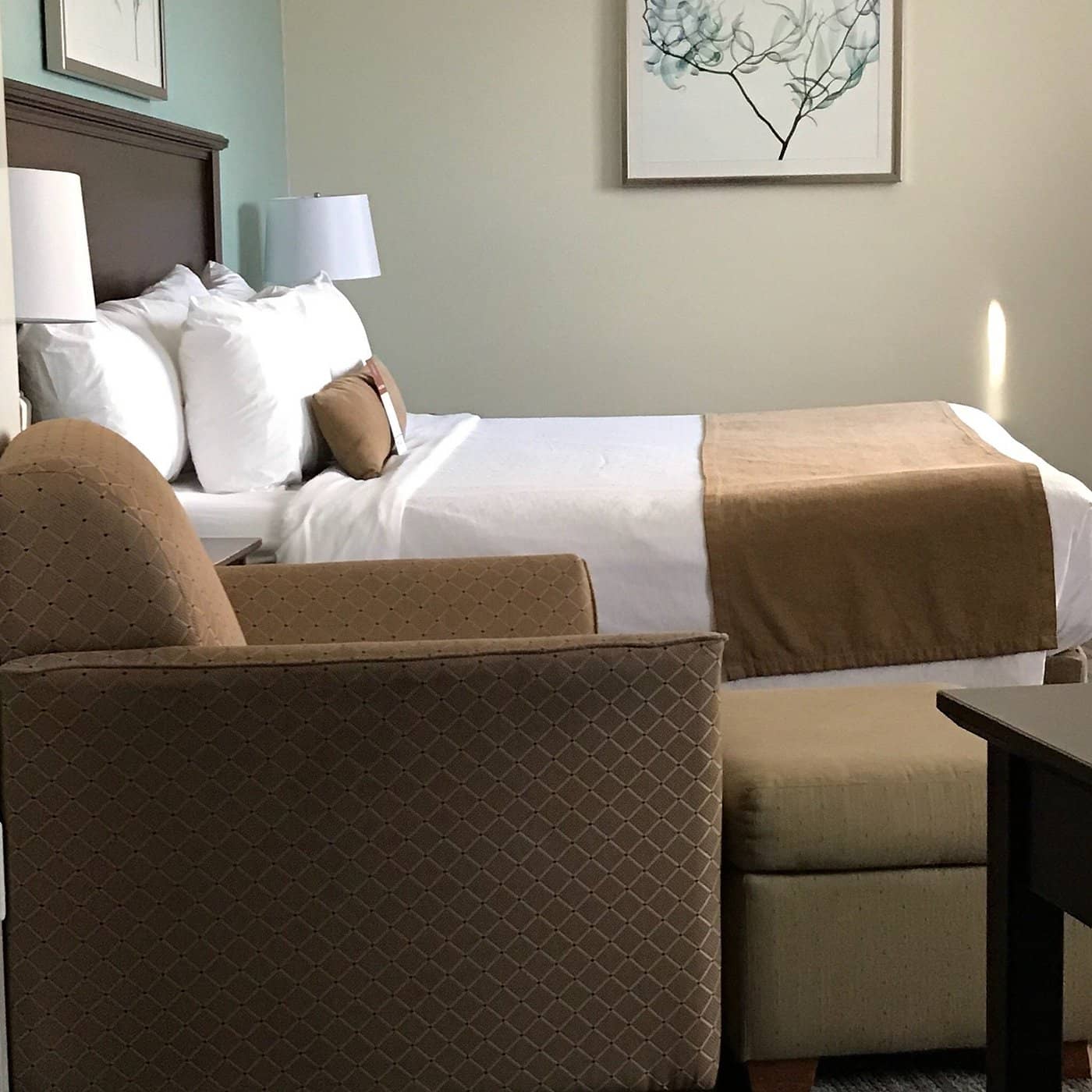 Cozy room at the Best Western Plus Chocolate Lake Hotel showcasing a neatly made queen-size bed with brown accents a comfy armchair a matching ottoman and simple decor featuring soft beige and light green tones