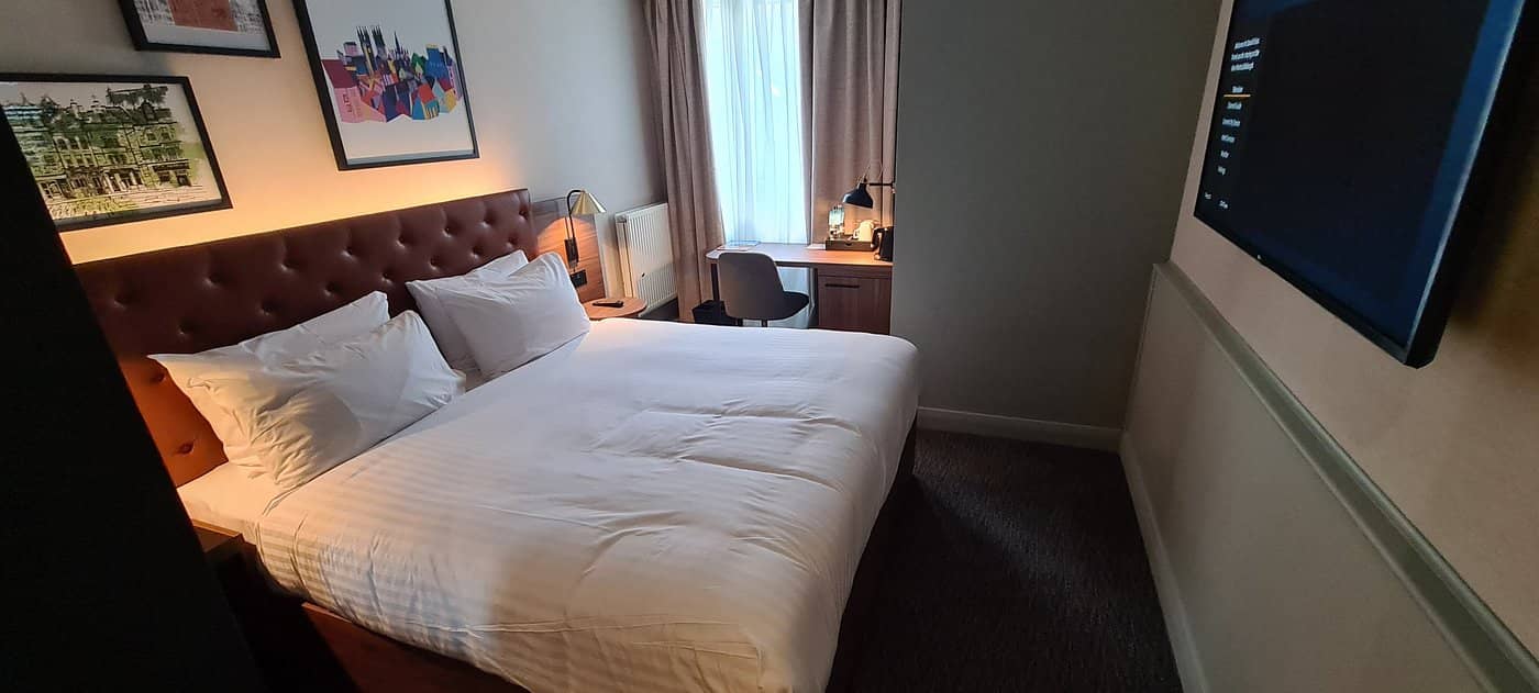 A cozy guest room at Four Points by Sheraton Edinburgh featuring a comfortable double bed with a tufted headboard soft lighting a well-equipped desk area and wall artwork adding a touch of elegance The room offers a warm and inviting ambiance perfect for a relaxing stay