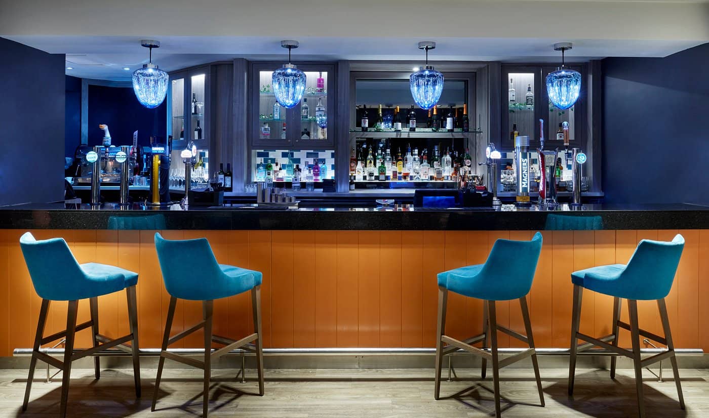 A stylish bar at Leonardo Edinburgh Murrayfield with an orange counter teal bar stools and a well-stocked liquor display