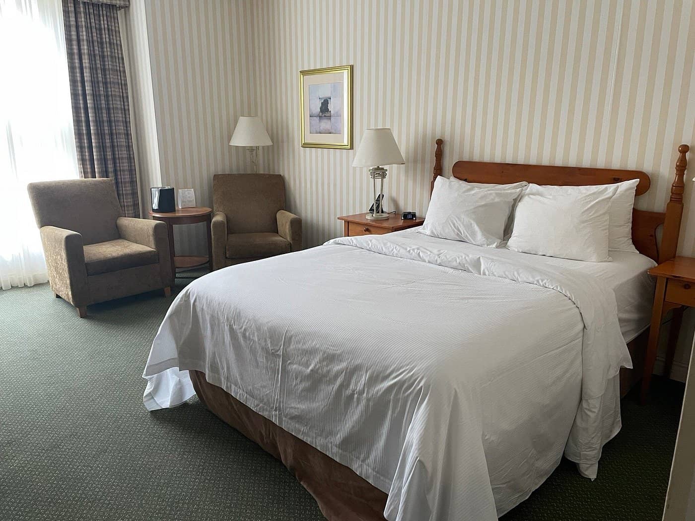 A guest room at The Barrington Hotel featuring a cozy queen-sized bed two comfortable chairs by the window and warm traditional decor with a wood headboard and matching side tables