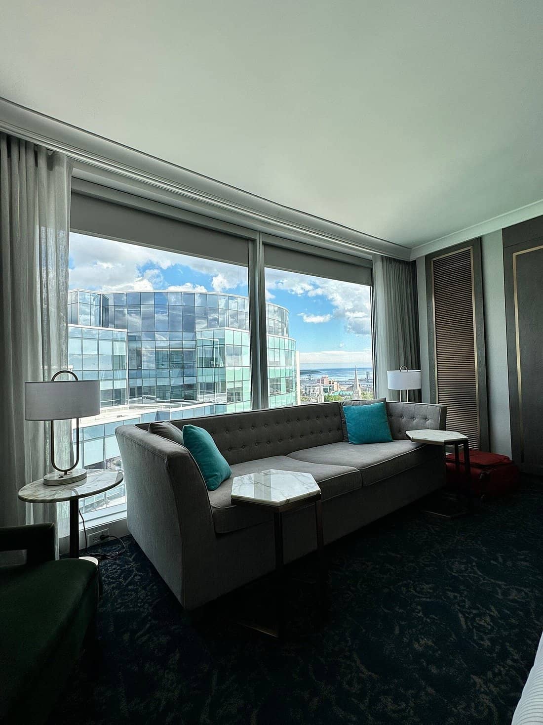 Modern suite at The Sutton Place Hotel Halifax featuring a sleek sofa with blue accent pillows two marble side tables and large windows offering a panoramic view of the city skyline and waterfront