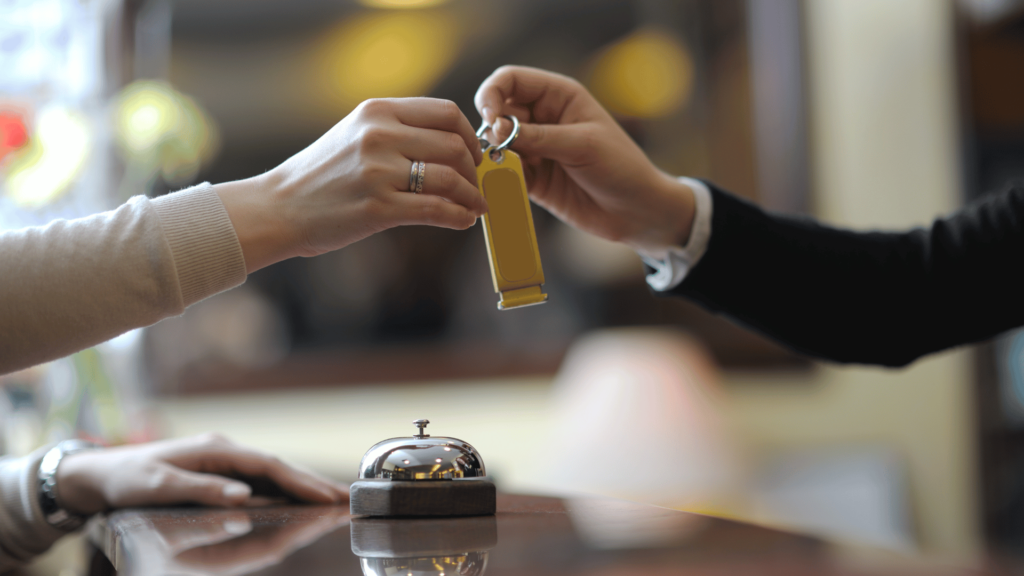 Close-up of a guest receiving a key from a receptionist at a hotel front desk, with a bell in the foreground. The warm, blurred background creates an inviting atmosphere, representing a friendly check-in experience at a budget hotel.