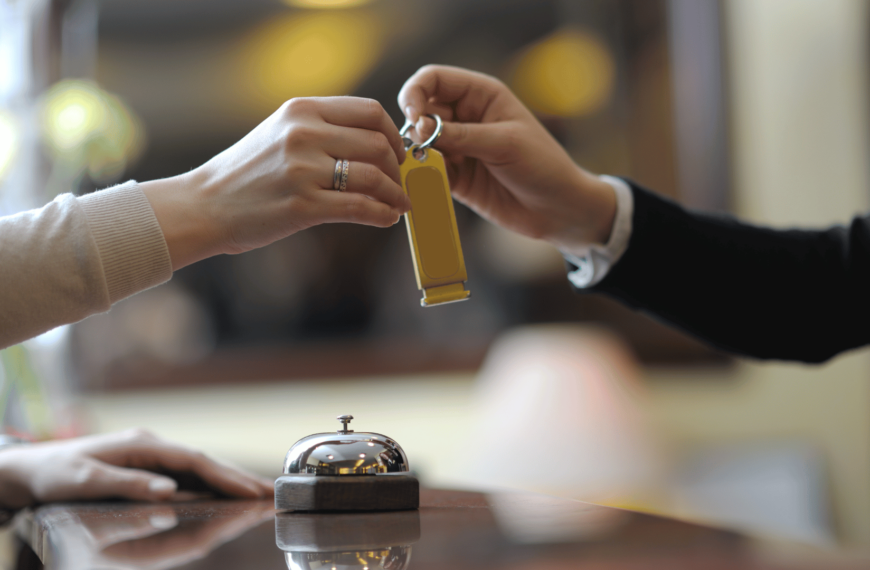 Close-up of a guest receiving a key from a receptionist at a hotel front desk, with a bell in the foreground. The warm, blurred background creates an inviting atmosphere, representing a friendly check-in experience at a budget hotel.