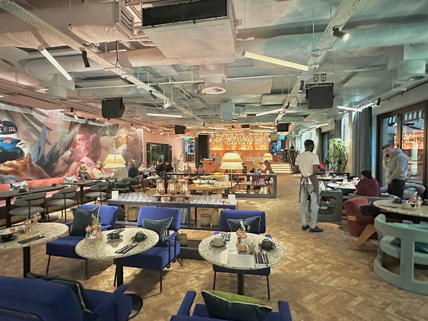 Dining area at Virgin Hotels Edinburgh showcasing a spacious modern interior with artistic wall murals vibrant blue seating round tables and warm ambient lighting Guests are seen enjoying their meals while staff members move around attentively