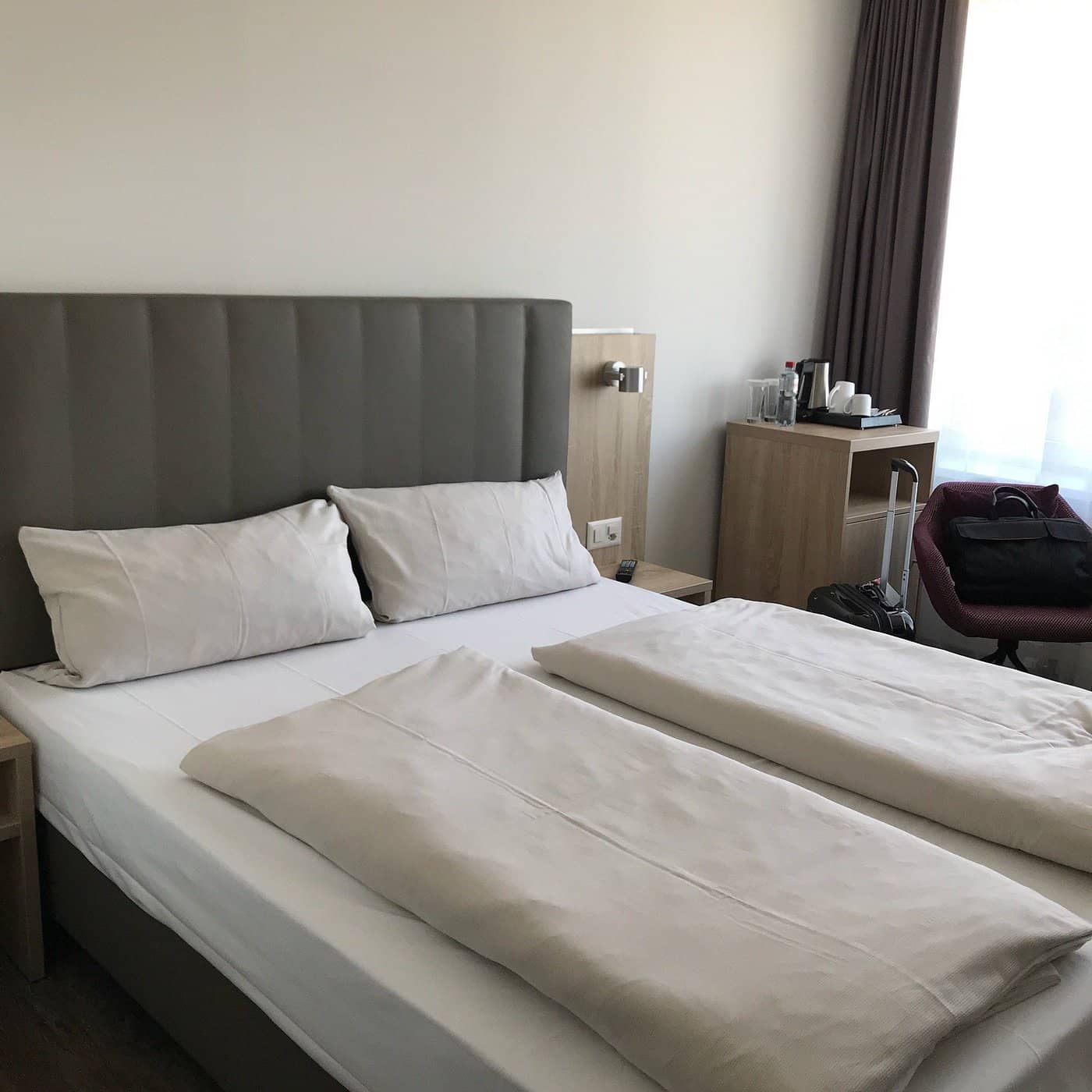 Modern room at Essential by Dorint Basel City featuring a comfortable bed with soft linens a padded headboard a small side table and a refreshment station with bottled water and cups creating a cozy and functional space