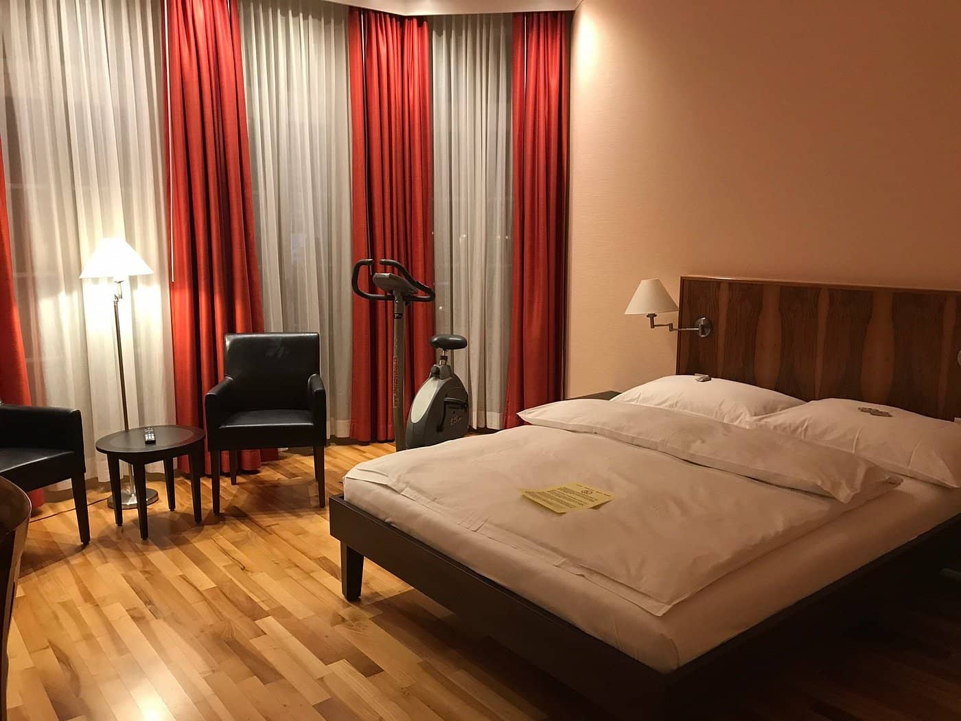 Spacious room at Hotel Schweizerhof Basel featuring a cozy bed seating area exercise bike and warm red curtains