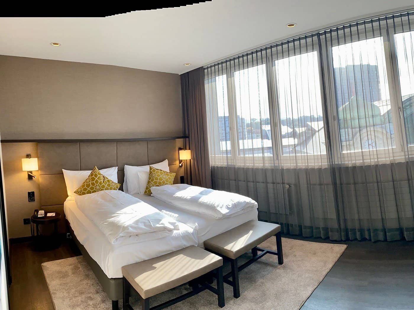 Modern sunlit hotel room at Hotel Victoria with twin beds yellow accent pillows bedside lamps and large windows with sheer curtains