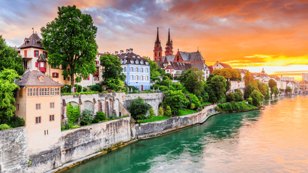 Scenic view of Basel, Switzerland at sunset, featuring historic buildings along the Rhine River with vibrant architecture and lush greenery.