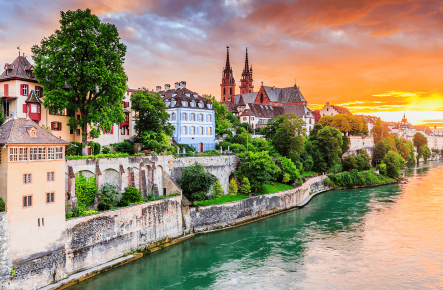 Scenic view of Basel, Switzerland at sunset, featuring historic buildings along the Rhine River with vibrant architecture and lush greenery.