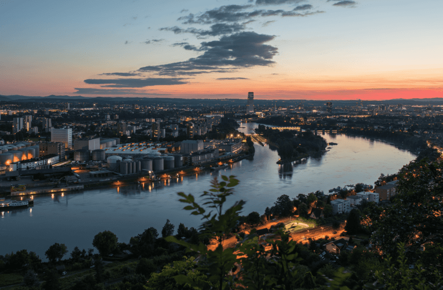 Scenic evening view of Basel, Switzerland, with city lights reflecting on the Rhine River and a colorful sunset sky.
