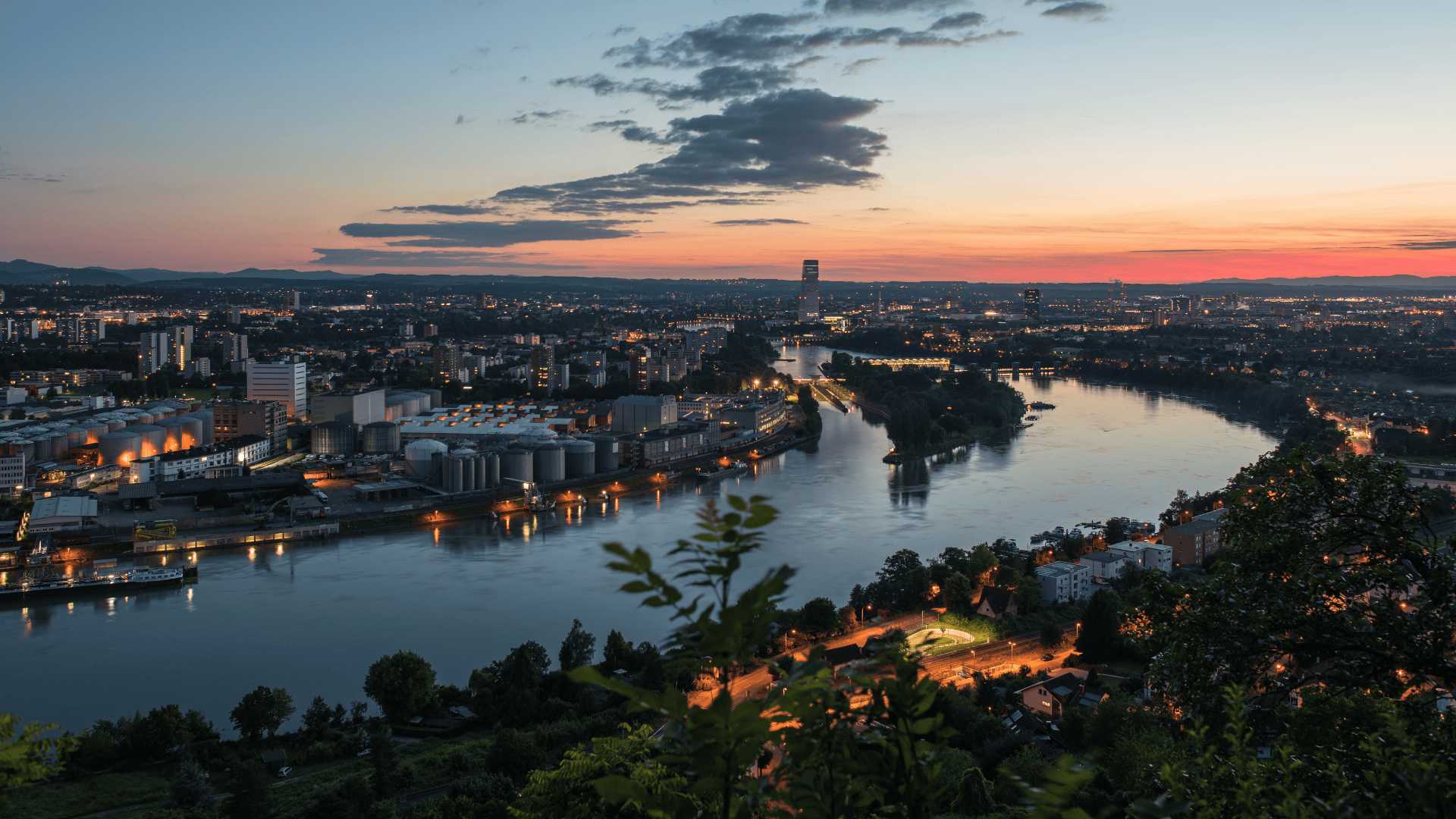 Scenic evening view of Basel, Switzerland, with city lights reflecting on the Rhine River and a colorful sunset sky.