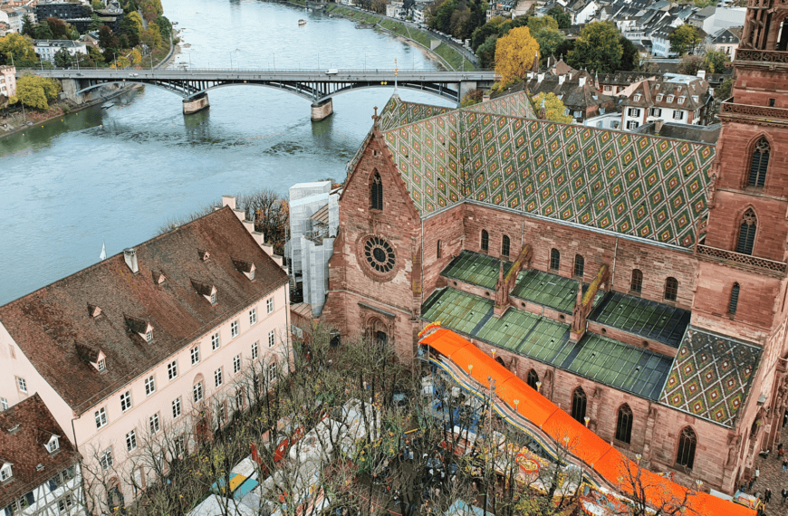 Aerial view of Basel Minster with its colorful tiled roof, situated near the Rhine River and surrounded by historic buildings and trees.