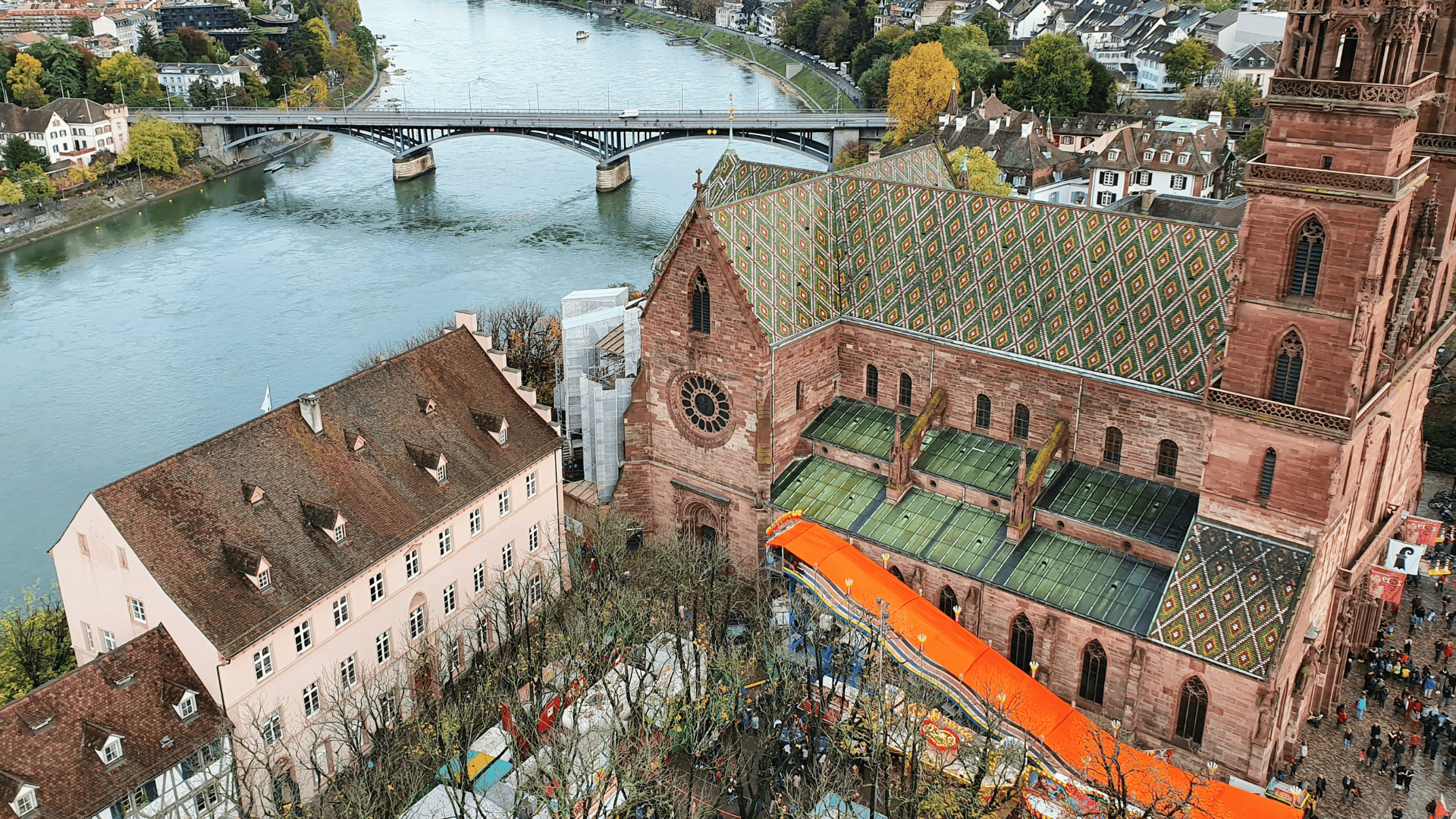 Aerial view of Basel Minster with its colorful tiled roof, situated near the Rhine River and surrounded by historic buildings and trees.