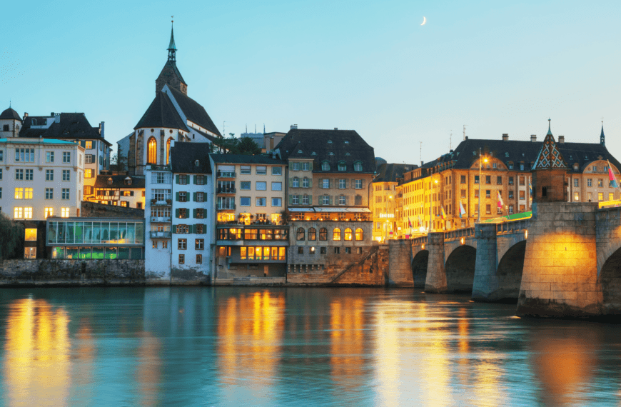 Evening view of Basel's Old Town with historic buildings and illuminated bridge reflecting on the Rhine River.