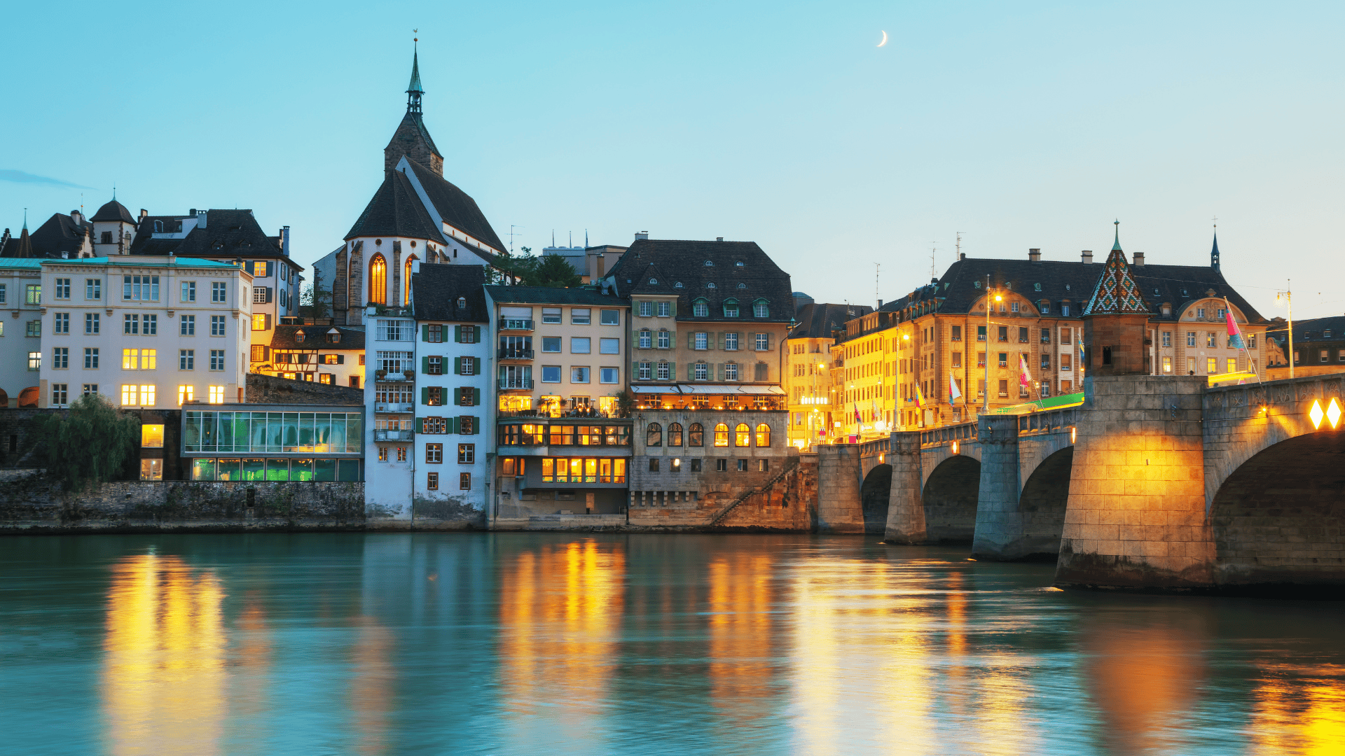 Evening view of Basel's Old Town with historic buildings and illuminated bridge reflecting on the Rhine River.